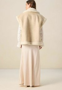 Femme portant un gilet sans manches en fausse fourrure beige par-dessus un haut à manches longues en dentelle blanche et une longue jupe crème, se tenant contre un fond uni.