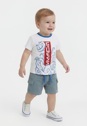 Dijete koje hoda, nosi bijelu majicu s plavim hobotnicom i crvenim okomitim slovima, sive cargo kratke hlače i bijele tenisice.