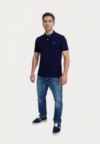 Man stands confidently in a navy polo shirt and blue jeans, with casual sneakers, against a light background.