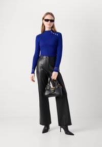 Royalblå ribbstickad tröja med knappar i sidan, svarta vida byxor i läder, svart handväska med gulddetaljer och svarta spetsiga klackskor.