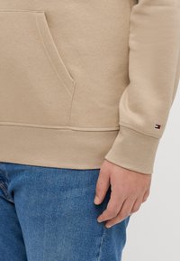 Beige sweatshirt with a kangaroo pocket, ribbed cuffs, and hem. Features a small logo accent on the sleeve. Paired with blue jeans.