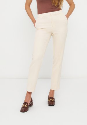 Γυναίκα που φοράει εκρού παντελόνι μέχρι τον αστράγαλο, καφέ αμάνικο τοπ και καφέ μοκασίνια με χρυσή αγκράφα, στέκεται με τα χέρια στις τσέπες.