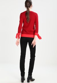 Suéter de punto rojo con mangas tres cuartos que presenta puños rojos contrastantes atados en lazo, combinado con jeans negros ajustados y botas de tacón.