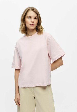 Vrouw met schouderlang blond haar, die een los lichtroze T-shirt en beige broek draagt, staat met één hand achter haar rug.