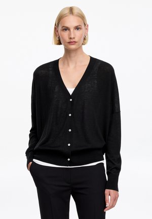 Femme aux cheveux blonds courts portant un cardigan noir boutonné sur un haut blanc et un pantalon noir, mains dans les poches, fond neutre.