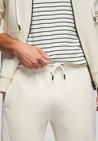 Homme portant un sweat à capuche crème zippé, un t-shirt rayé noir et blanc, et un pantalon de jogging crème à cordon de serrage, ajustant la taille du t-shirt.