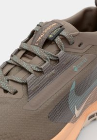 Brown athletic shoe featuring a textured upper, turquoise laces, GORE-TEX branding, and an orange outsole. Detailed with a patterned design.