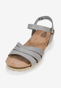 Sandalia de cuero gris con tres tiras cruzadas, correa para el tobillo ajustable y una plantilla texturada marrón. Presenta una suela flexible de goma color crema.