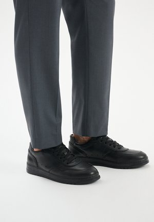 Czarne skórzane sneakersy z okrągłym noskiem, teksturowaną powierzchnią i perforowanymi detalami. Noszone z szarymi, dopasowanymi spodniami. Niski krój.