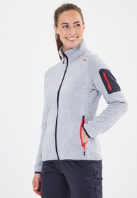 Graue Zip-Jacke mit hohem Kragen, roten Akzenten an Reißverschlüssen und Taschen sowie strukturiertem Stoff. Hat einen schwarzen Bereich am linken Ärmel.