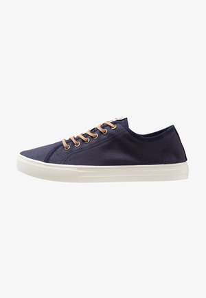 Zapatilla deportiva baja azul marino con cordones beige y suela de goma blanca, vista desde el lado exterior sobre fondo blanco.