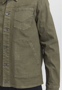 Olivnozeleni jeans jakna z gumbom spredaj, žepom na prsih in stranskimi žepi; ima strukturiran dizajn in gladko teksturo.