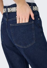 Jean en denim bleu foncé avec une coupe ajustée. Une ceinture texturée noire et blanche à motif serpent, avec une main placée dans la poche arrière.