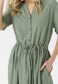 Grön klänning med prickar, tillverkad av lättviktig tyg, med knäppning fram, korta ärmar, elastisk midja och sidfickor.