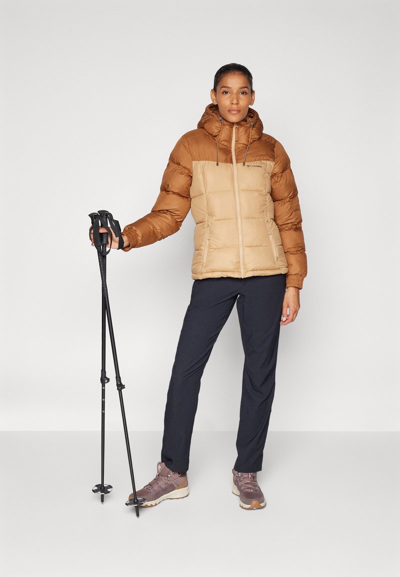 Brązowa i beżowa izolowana kurtka puchowa z kapturem, z zamykanymi kieszeniami, noszona z ciemnymi spodniami i butami trekkingowymi, trzymająca kijki trekkingowe.