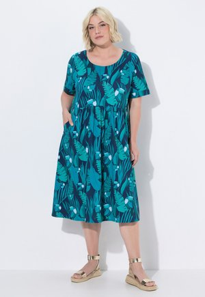 Vrouw met kort blond haar die een zeegroene jurk draagt met groene tropische bladeren en vogelprint, gouden sleehak sandalen, staand tegen een witte achtergrond.