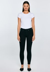 Sieviete stāv, tērpusies piegulošā baltā īsā piedurkņu topā, melnās augsta vidukļa saspringtās džinsās un melnās punktētas kurpēs plakanoj fona priekšā.