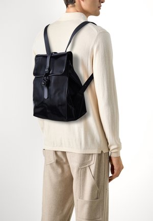 Zaino nero in nylon con patta pieghevole, spallacci regolabili e design elegante, caratterizzato da una fibbia frontale prominente e un brand discreto.