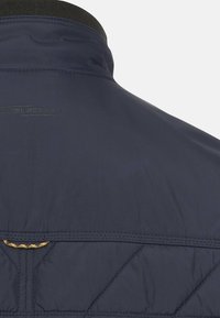Veste légère bleu marine avec un col montant, une texture matelassée et un petit logo au dos. Présente un détail de surpiqûres jaunes en accent.