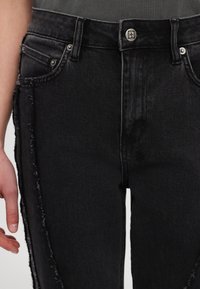 Jean en denim noir avec coutures latérales effilochées et fermeture par bouton argenté, porté avec un haut sombre rentré et une main gauche visible.