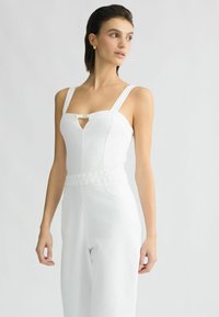Combinaison blanche avec décolleté carré, bretelles larges et ceinture tressée. Présente une ouverture en forme de clé à l'avant et un tissu lisse et texturé.