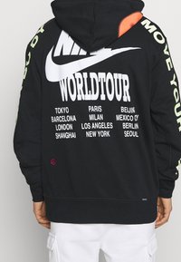 Svart hoodie med stor vit "Nike"-logotyp och texten "WORLD TOUR". Innehåller stadnamn i vitt, neongröna accenter och rosa logodetalj.