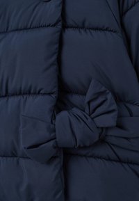 Doudoune bleu marine avec un détail de nœud noué ; possède des sections matelassées et une texture douce et lisse. Fermeture à boutons sur le devant.