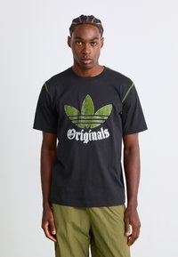 Fekete pamut póló, amelyen egy megkopott zöld Adidas trefoil logó és fehér "Originals" felirat található, zöld díszvarrással a vállakon.