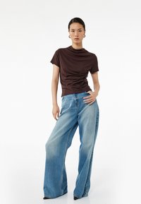 Brun tætsiddende t-shirt med samledetalje på den ene side, parret med lyseblå vidde jeans med en falmet struktur og store lommer.