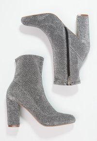 Botines de brillo gris con un alto tacón cuadrado, que cuentan con una cremallera lateral y una punta afilada. La textura brilla bajo la luz.