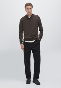 Maglione lavorato a maglia marrone con colletto alto, tre bottoni e polsini a costine; abbinato a pantaloni neri larghi e scarpe scure. Sfondo neutro.