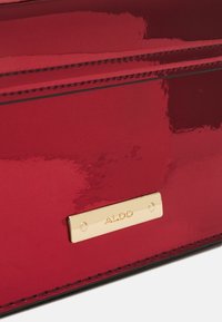 Sac à main rouge brillant avec coutures noires et plaque logo ALDO en métal doré. Texture lisse avec un design structuré et des lignes épurées.