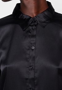 Camicia nera in satin con colletto a punta, chiusura con bottoni e superficie liscia. Presenta una finitura lucida e un leggero riflesso.
