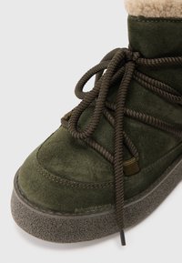 Stivale verde in suede con suola in gomma testurizzata. Presenta lacci marroni e un morbido colletto in pile crema. Inquadratura ravvicinata del lato anteriore.