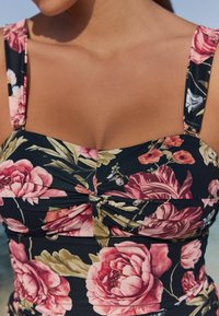 Costume da bagno floreale con sfondo nero, caratterizzato da grandi peonie rosa e foglie verdi. Design arricciato e spalline spesse.