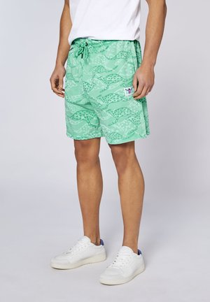 Mann trägt mintgrüne Shorts mit abstraktem Gepardmuster, weiße Sneakers und ein weißes T-Shirt vor einfachem Hintergrund.