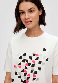 White short-sleeve t-shirt features a geometric pattern of black, pink, grey, and cream semicircles. Smooth texture and round neckline.