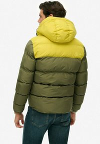Chaqueta acolchada que presenta una parte superior amarilla y una sección inferior en verde olivo. Tiene capucha, textura acolchada horizontal y un logo en la parte trasera.