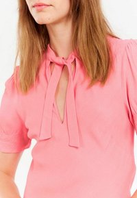 Blouse rose avec de courtes manches bouffantes, détail à nouer au niveau du col et décolleté en V. Texture de tissu lisse et design simple. Pas de motifs ni de matériel.