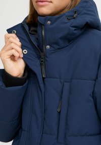 Navy blue insulated jacket with a high collar, visible zippers, snap buttons, and a side pocket. Slightly glossy finish, quilted texture.