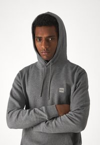 Grijze hoodie van zacht materiaal, met een voorzak en een logopatch. Het heeft een capuchon met trekkoord en ribbels aan de mouwen.