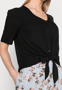 Top nero a maniche corte con scollatura a V, caratterizzato da una chiusura con bottoni davanti e un dettaglio annodato in vita. Tessuto morbido con una texture liscia.