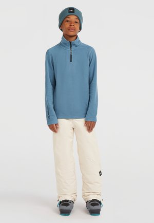 Blauwe fleece longsleeve met rits, crèmekleurige sneeuwbroeken en grijze sneeuwlaarzen met teal-accenten, gedragen met een bijpassende blauwe beanie.