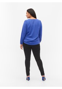 Top azul texturizado de manga larga con escote redondeado y una pequeña abertura en la parte trasera, combinado con pantalones ajustados negros y sandalias de tacón azules.