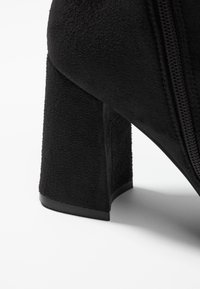 Bota negra de ante hasta el tobillo con un tacón grueso, que presenta una cremallera lateral y una superficie suave y texturizada.