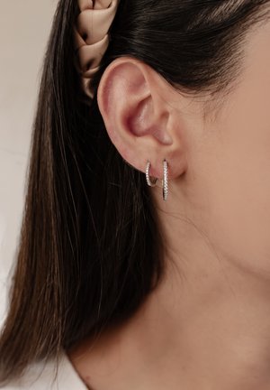 Silberne Creolen mit strukturierter Oberfläche, die mit kleinen klaren Edelsteinakzenten versehen sind. Getragen an einem menschlichen Ohr, wobei dunkles Haar teilweise sichtbar ist.