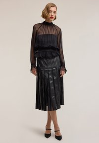 Donna con capelli biondi corti che indossa una camicetta nera trasparente a maniche lunghe e una gonna midi plissettata in pelle nera con tacchi neri, su sfondo semplice.