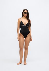 Juodas vientisas maudymosi kostiumėlis su plonais reguliuojamais dirželiais ir gilia iškirpte. Ant krūtinės kerta kontrastinga plona horizontali juosta.