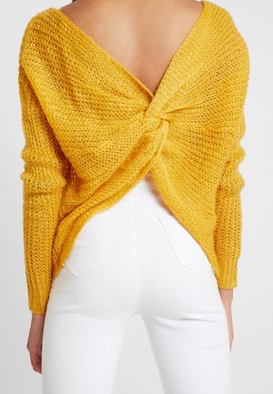 Vue de dos d'une personne portant un pull jaune en maille avec un dos ouvert torsadé et un pantalon blanc taille haute.