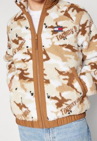 Giacca in pile con motivo mimetico beige, zip e rifiniture marroni, polsini elasticizzati e logo "Tommy Jeans" in evidenza sulla tasca del petto.